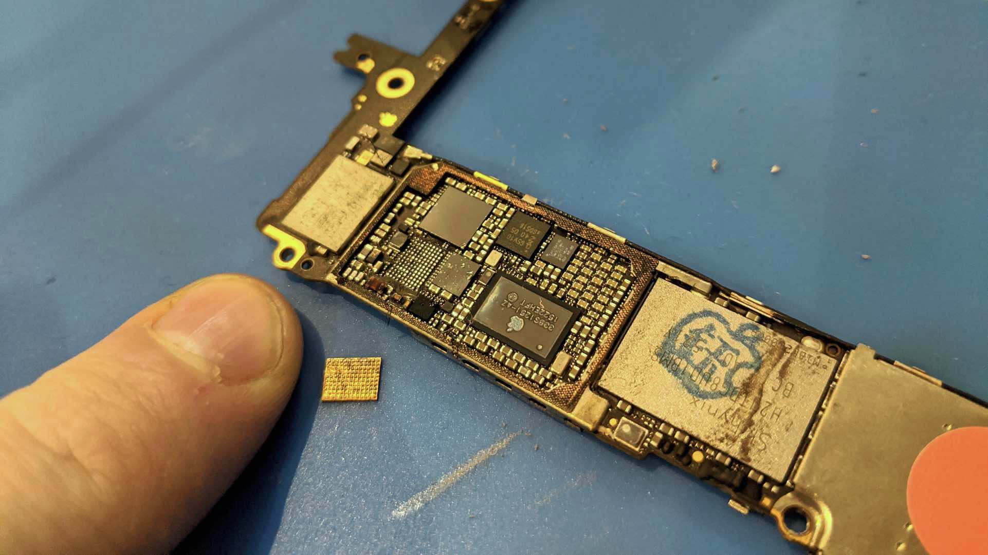 iPhone board with chip removed, next to fingernail for scale